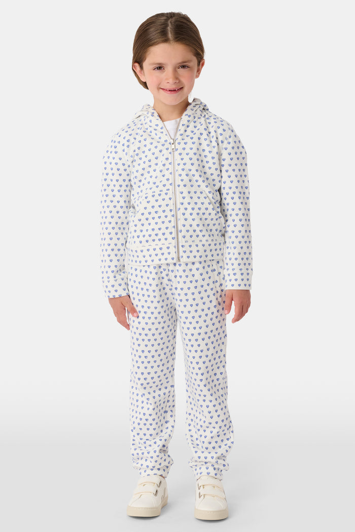 A smiling young child with brown hair wears the Roller Rabbit Kids Hearts Harley Hoodie and matching pants, both made from 100 cotton French terry, standing with hands in the hoodie’s pockets against a plain light background.