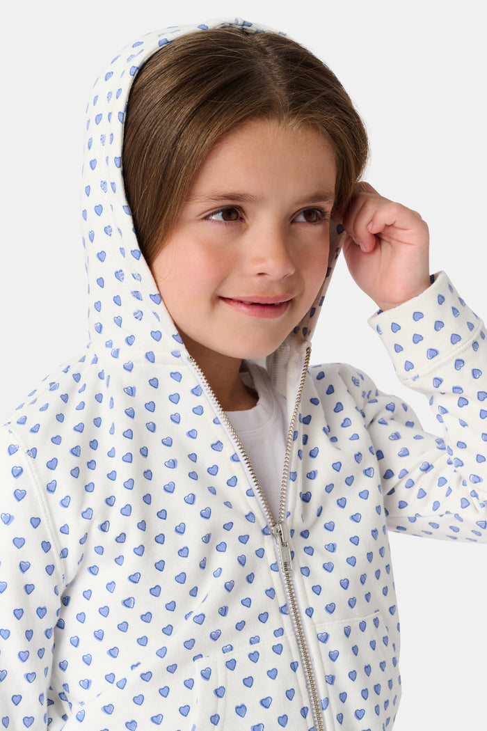 A smiling young child with brown hair wears the Roller Rabbit Kids Hearts Harley Hoodie and matching pants, both made from 100 cotton French terry, standing with hands in the hoodie’s pockets against a plain light background.
