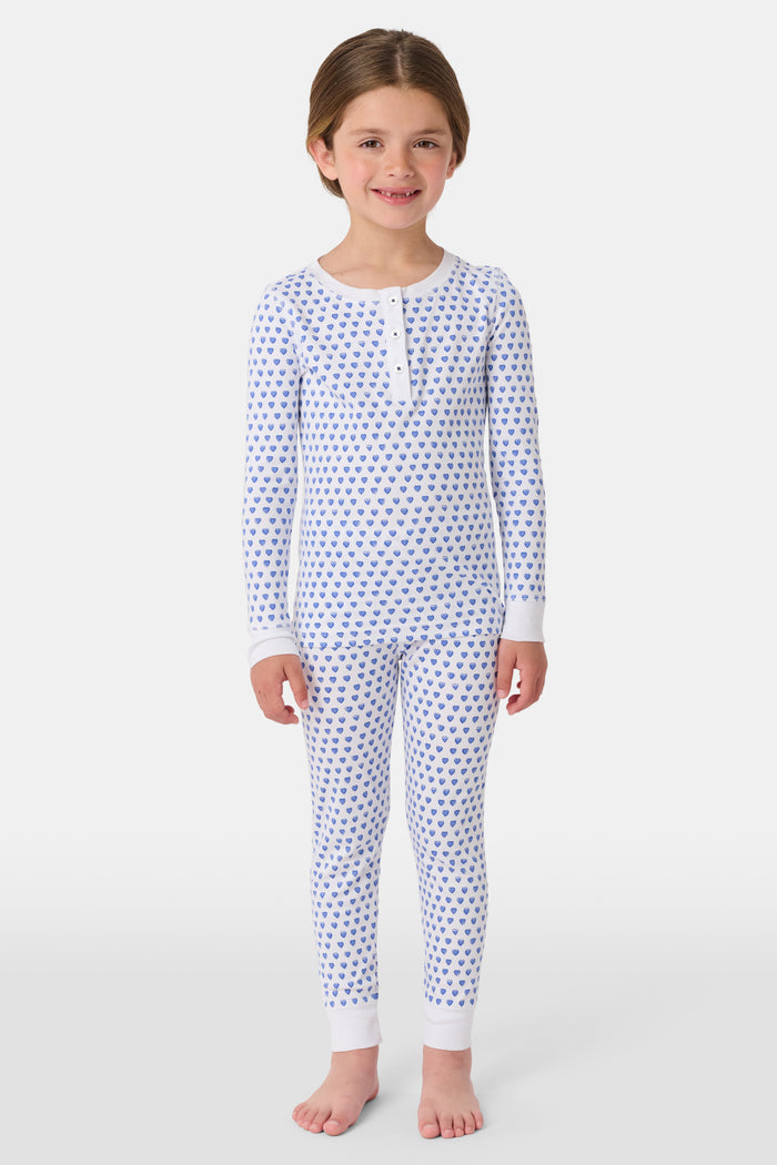 A girl smiles barefoot in Roller Rabbit’s Kids Hearts Pajamas, a matching set featuring a heart print, and poses against a plain white background.