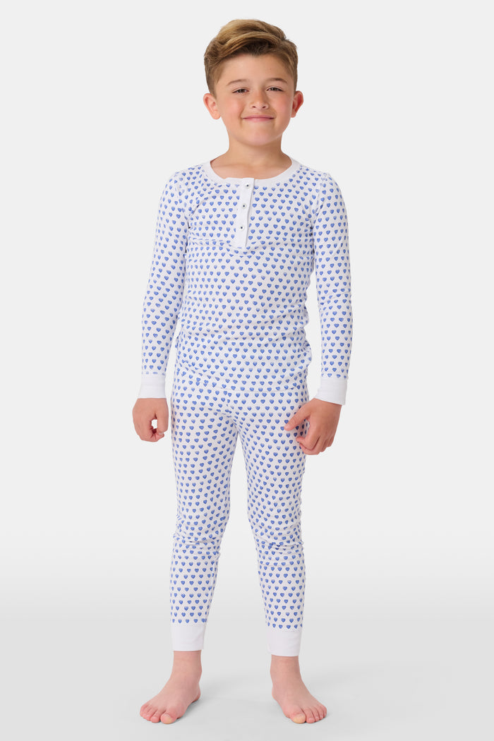 A girl smiles barefoot in Roller Rabbit’s Kids Hearts Pajamas, a matching set featuring a heart print, and poses against a plain white background.