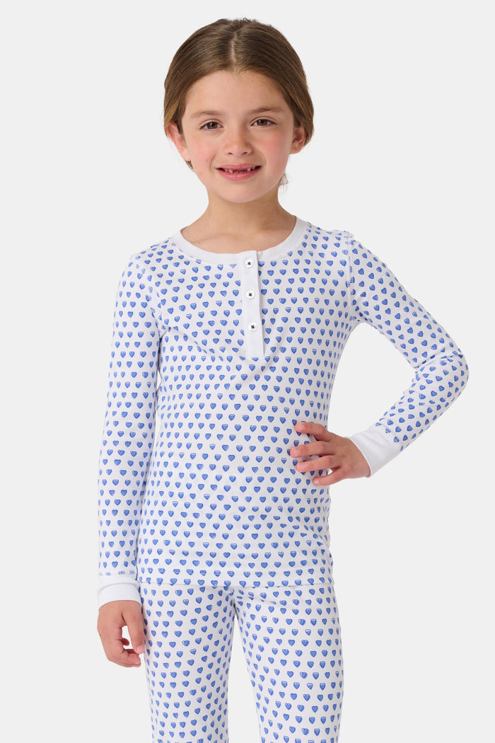 A girl smiles barefoot in Roller Rabbit’s Kids Hearts Pajamas, a matching set featuring a heart print, and poses against a plain white background.