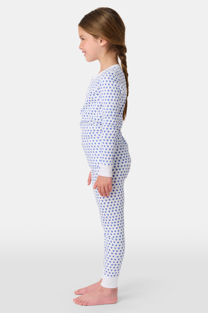 A girl smiles barefoot in Roller Rabbit’s Kids Hearts Pajamas, a matching set featuring a heart print, and poses against a plain white background.