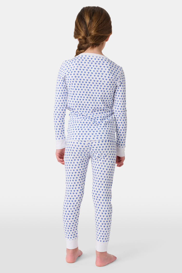 A girl smiles barefoot in Roller Rabbit’s Kids Hearts Pajamas, a matching set featuring a heart print, and poses against a plain white background.