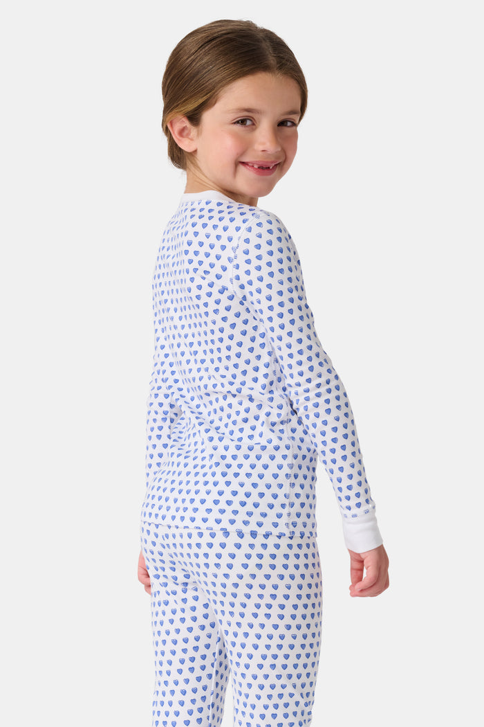 A girl smiles barefoot in Roller Rabbit’s Kids Hearts Pajamas, a matching set featuring a heart print, and poses against a plain white background.