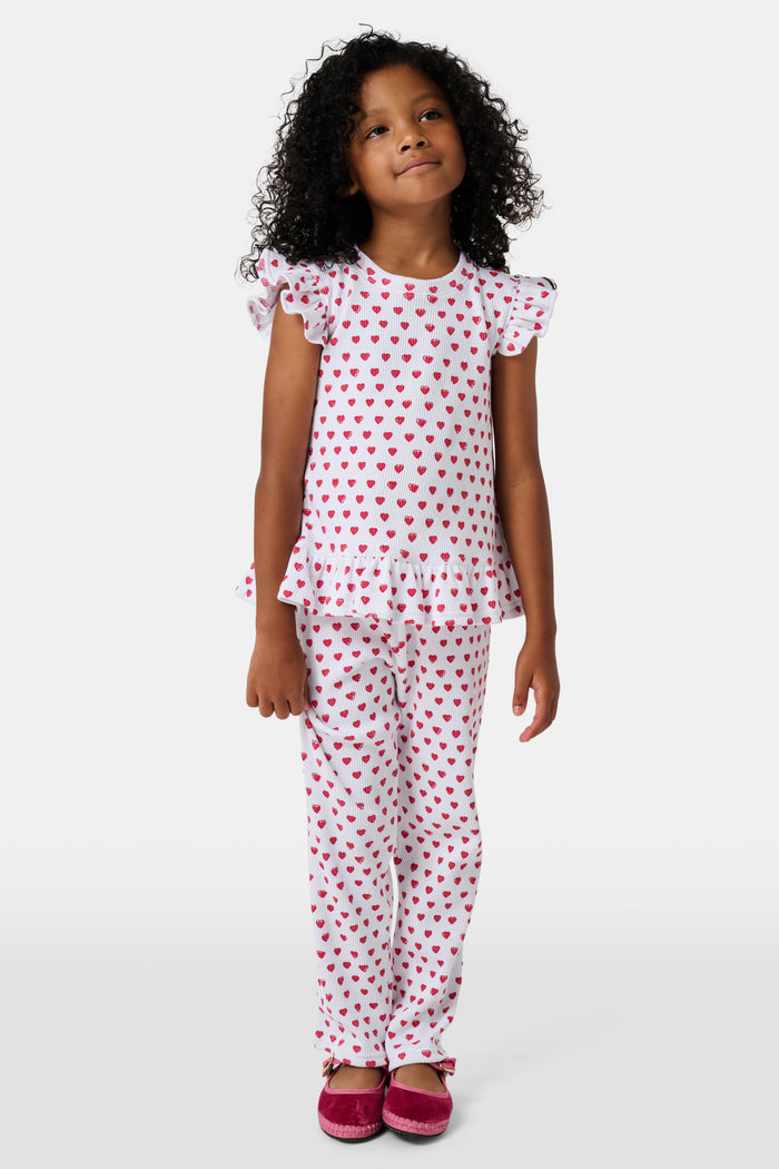 A child with long blonde hair wears the Roller Rabbit Kids Hearts Riley Set made from Pima Waffle fabric, featuring red hearts and ruffled cap sleeves, while holding a notebook with a decorative cover and standing in front of lockers.