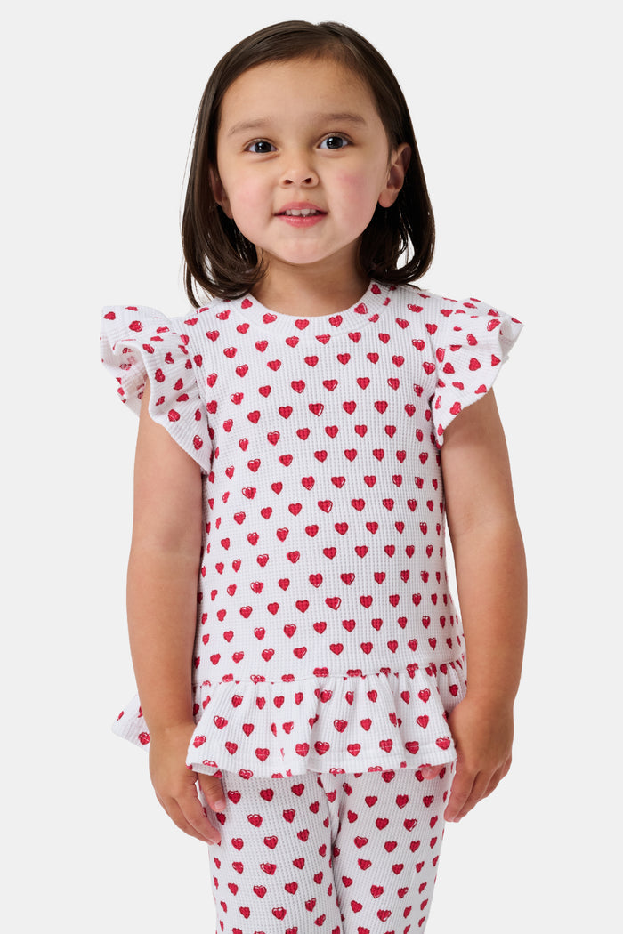 A child with long blonde hair wears the Roller Rabbit Kids Hearts Riley Set made from Pima Waffle fabric, featuring red hearts and ruffled cap sleeves, while holding a notebook with a decorative cover and standing in front of lockers.