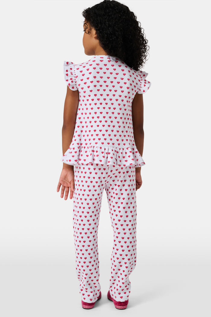 A child with long blonde hair wears the Roller Rabbit Kids Hearts Riley Set made from Pima Waffle fabric, featuring red hearts and ruffled cap sleeves, while holding a notebook with a decorative cover and standing in front of lockers.