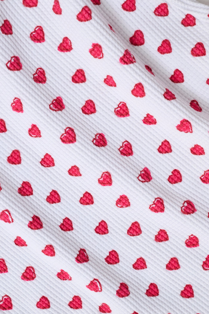 A child with long blonde hair wears the Roller Rabbit Kids Hearts Riley Set made from Pima Waffle fabric, featuring red hearts and ruffled cap sleeves, while holding a notebook with a decorative cover and standing in front of lockers.