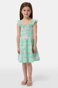 A young girl with straight brown hair smiles in the Roller Rabbit Kids Hydrangea Gingham Ceri Dress, featuring pink floral patterns and ruffled sleeves. She pairs it with white sandals and stands against a plain white background.