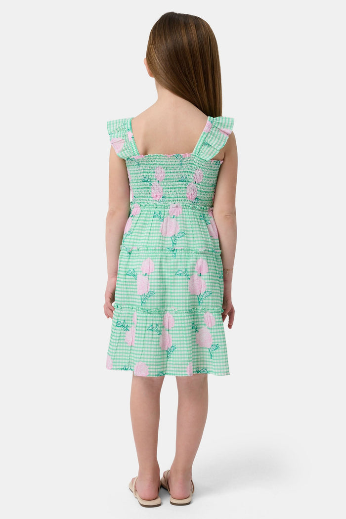 A young girl with straight brown hair smiles in the Roller Rabbit Kids Hydrangea Gingham Ceri Dress, featuring pink floral patterns and ruffled sleeves. She pairs it with white sandals and stands against a plain white background.