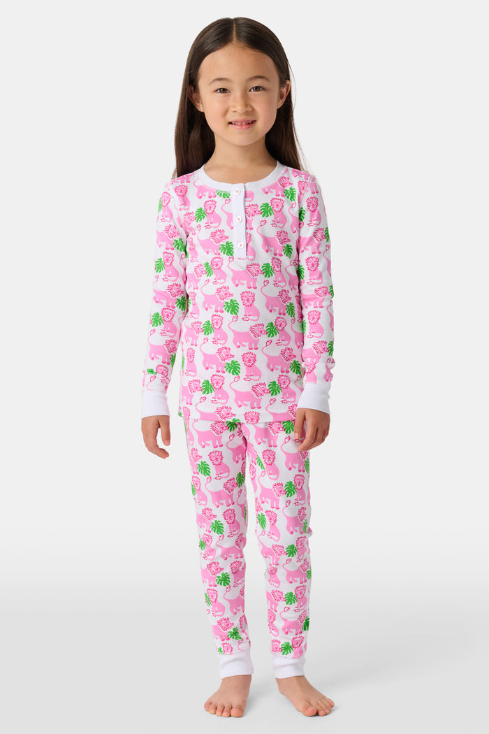 A young girl smiles barefoot against a white background in Roller Rabbit's Kids Leo The Lion Pajamas—soft Pima cotton, long sleeves, white cuffs, and a playful pink-and-green animal print.