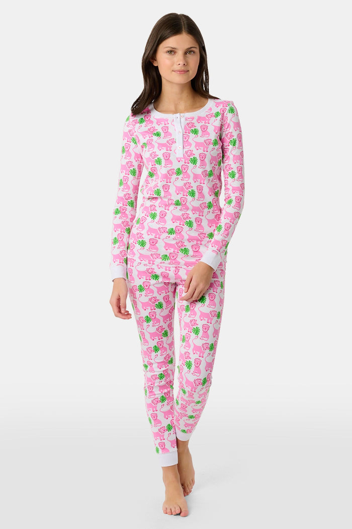 A young girl smiles barefoot against a white background in Roller Rabbit's Kids Leo The Lion Pajamas—soft Pima cotton, long sleeves, white cuffs, and a playful pink-and-green animal print.