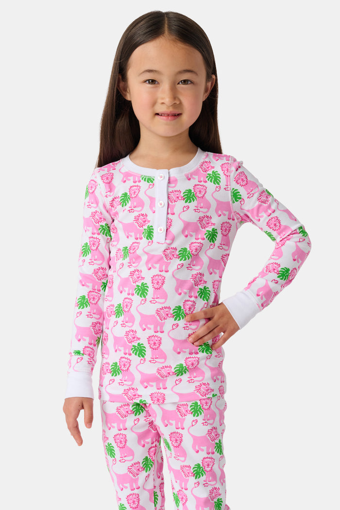 A young girl smiles barefoot against a white background in Roller Rabbit's Kids Leo The Lion Pajamas—soft Pima cotton, long sleeves, white cuffs, and a playful pink-and-green animal print.