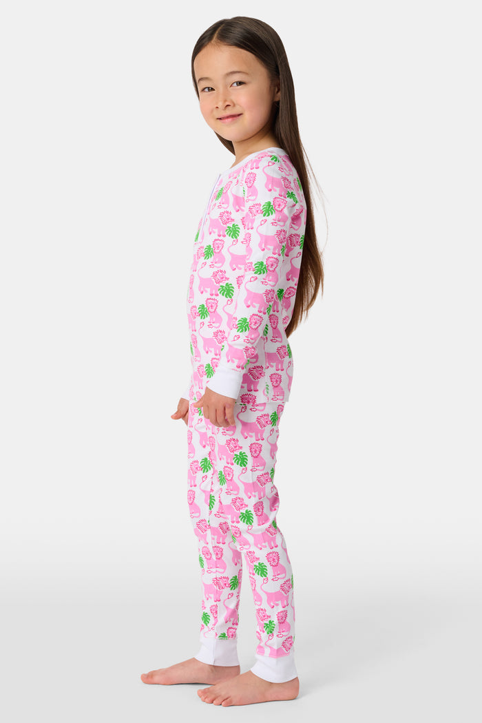 A young girl smiles barefoot against a white background in Roller Rabbit's Kids Leo The Lion Pajamas—soft Pima cotton, long sleeves, white cuffs, and a playful pink-and-green animal print.