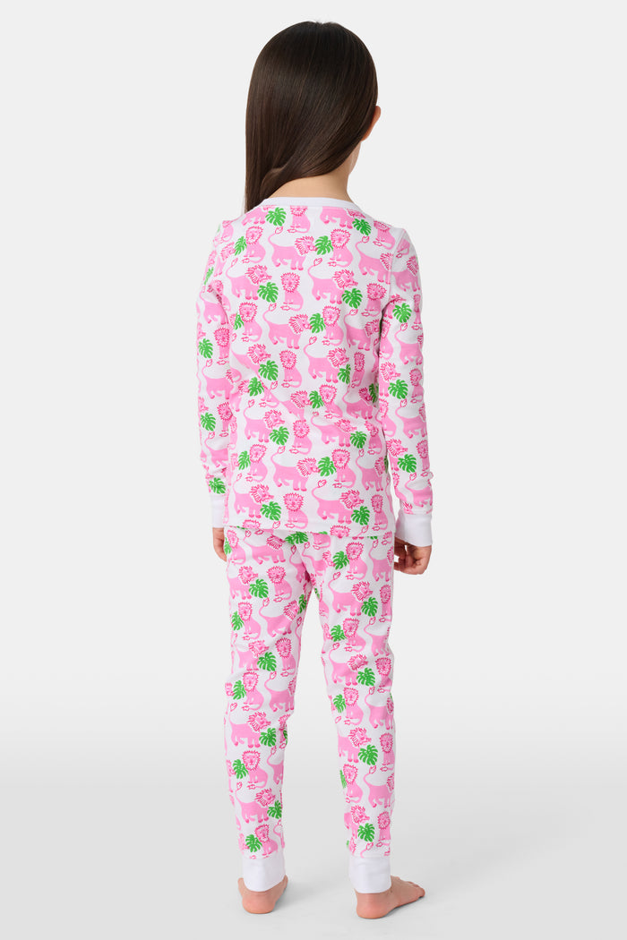 A young girl smiles barefoot against a white background in Roller Rabbit's Kids Leo The Lion Pajamas—soft Pima cotton, long sleeves, white cuffs, and a playful pink-and-green animal print.