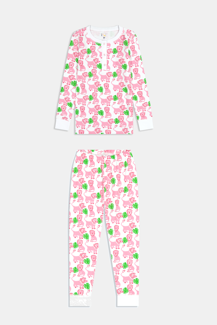 A young girl smiles barefoot against a white background in Roller Rabbit's Kids Leo The Lion Pajamas—soft Pima cotton, long sleeves, white cuffs, and a playful pink-and-green animal print.