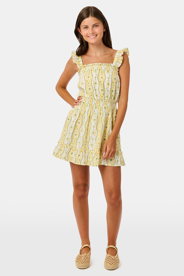 A girl wears the Roller Rabbit Kids Lillian Floral Stripe Tristan Dress, a cotton sundress with green and white stripes, floral patterns, ruffled straps, and a ruffled hem, standing with a smile and one arm relaxed at her side.