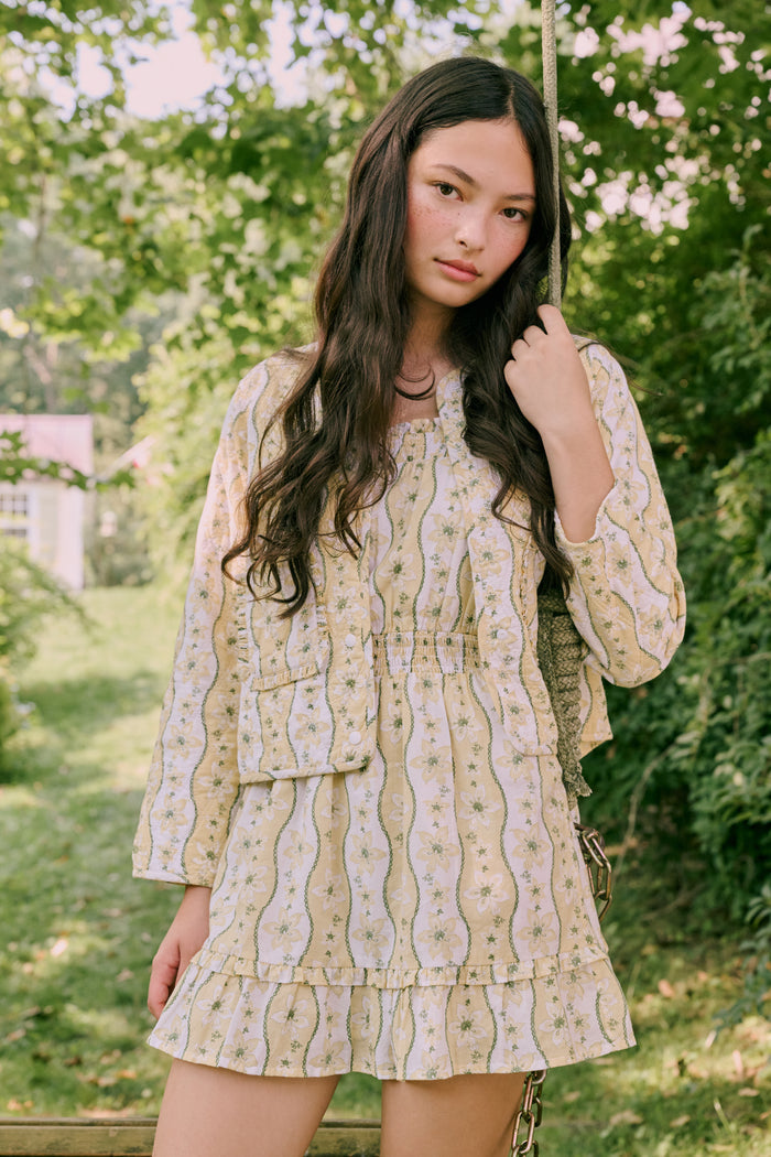 A girl wears the Roller Rabbit Kids Lillian Floral Stripe Tristan Dress, a cotton sundress with green and white stripes, floral patterns, ruffled straps, and a ruffled hem, standing with a smile and one arm relaxed at her side.