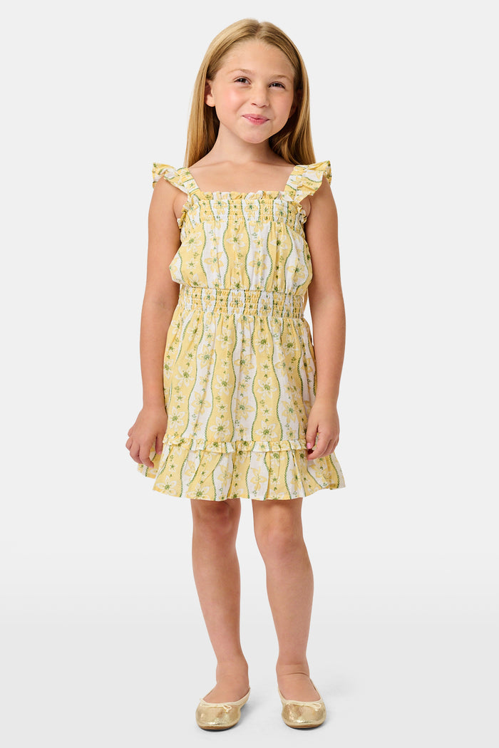 A girl wears the Roller Rabbit Kids Lillian Floral Stripe Tristan Dress, a cotton sundress with green and white stripes, floral patterns, ruffled straps, and a ruffled hem, standing with a smile and one arm relaxed at her side.