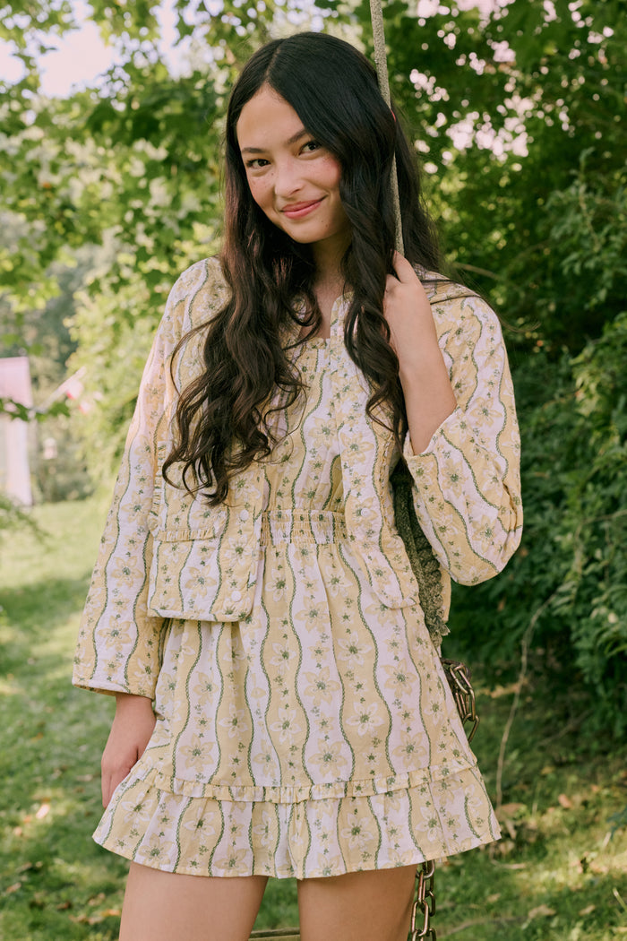 A girl wears the Roller Rabbit Kids Lillian Floral Stripe Tristan Dress, a cotton sundress with green and white stripes, floral patterns, ruffled straps, and a ruffled hem, standing with a smile and one arm relaxed at her side.