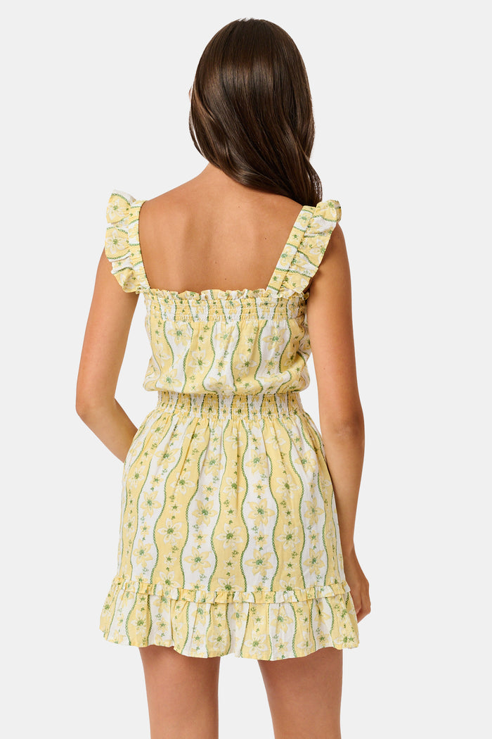 A girl wears the Roller Rabbit Kids Lillian Floral Stripe Tristan Dress, a cotton sundress with green and white stripes, floral patterns, ruffled straps, and a ruffled hem, standing with a smile and one arm relaxed at her side.