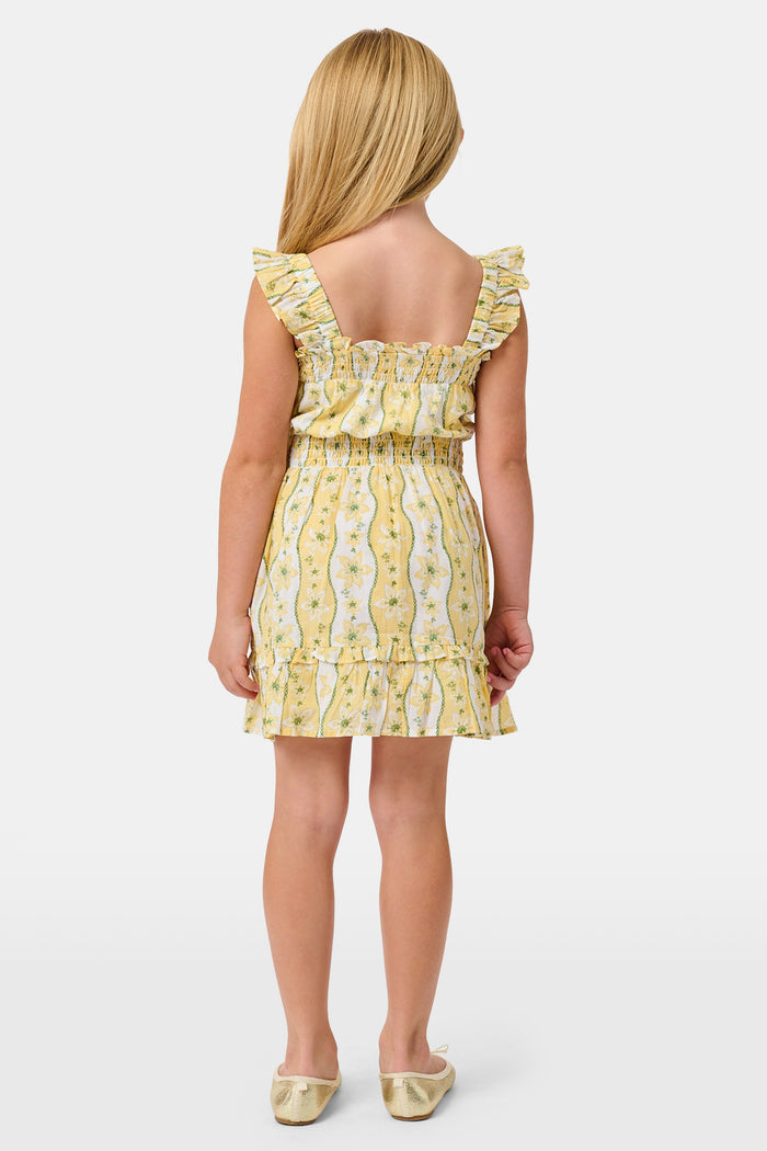 A girl wears the Roller Rabbit Kids Lillian Floral Stripe Tristan Dress, a cotton sundress with green and white stripes, floral patterns, ruffled straps, and a ruffled hem, standing with a smile and one arm relaxed at her side.