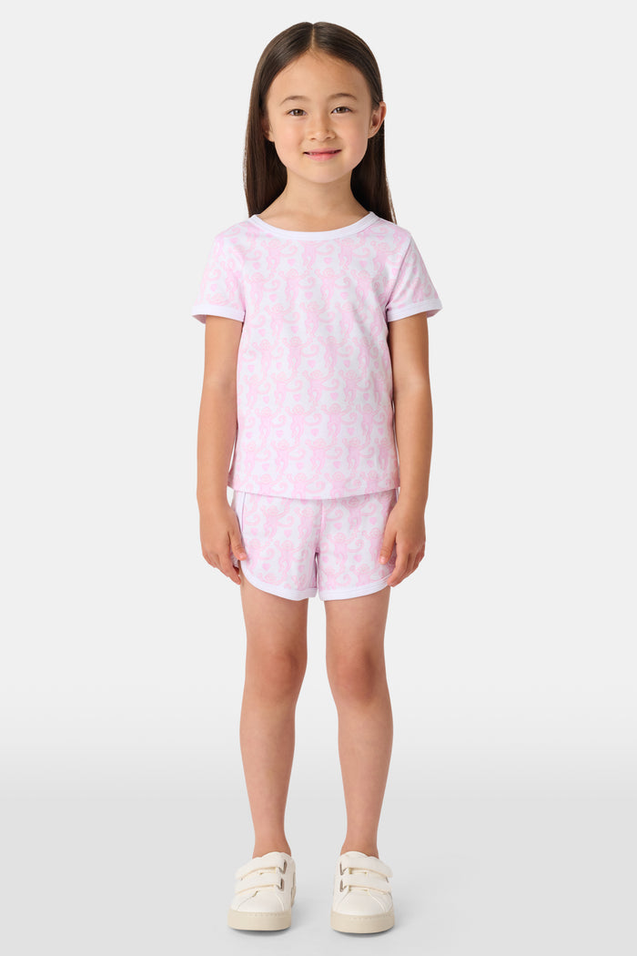 A young girl with straight brown hair wears the Roller Rabbit Kids Lovely Monkey Playa Play Set, a light pink Pima cotton pajama with short sleeves and shorts featuring a subtle monkey pattern as she smiles against a plain white background.