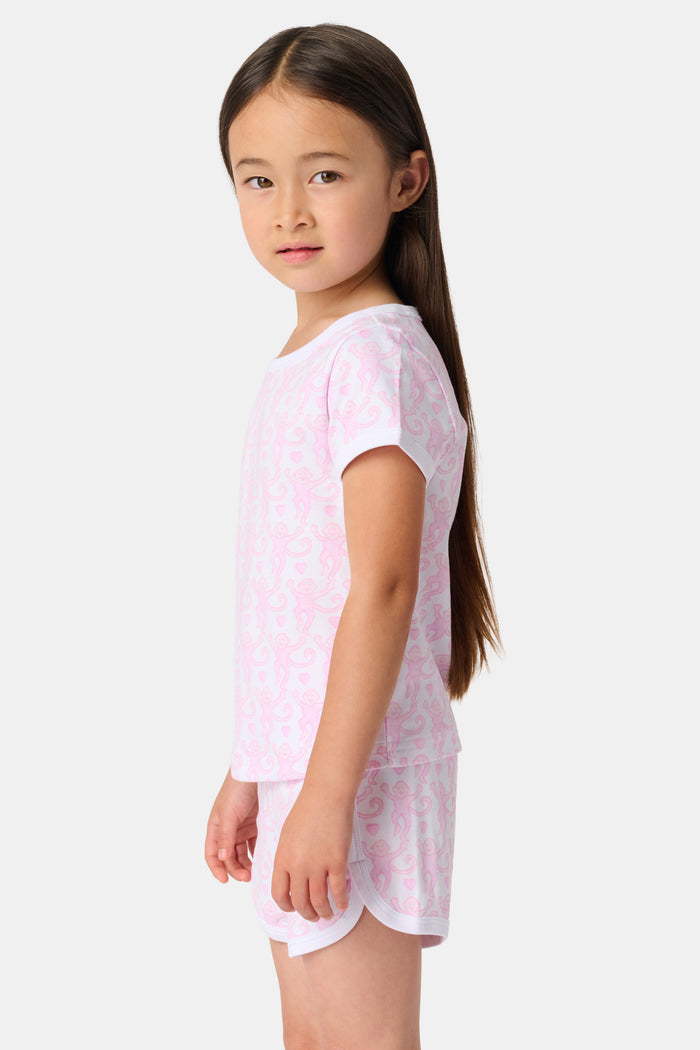 A young girl with straight brown hair wears the Roller Rabbit Kids Lovely Monkey Playa Play Set, a light pink Pima cotton pajama with short sleeves and shorts featuring a subtle monkey pattern as she smiles against a plain white background.