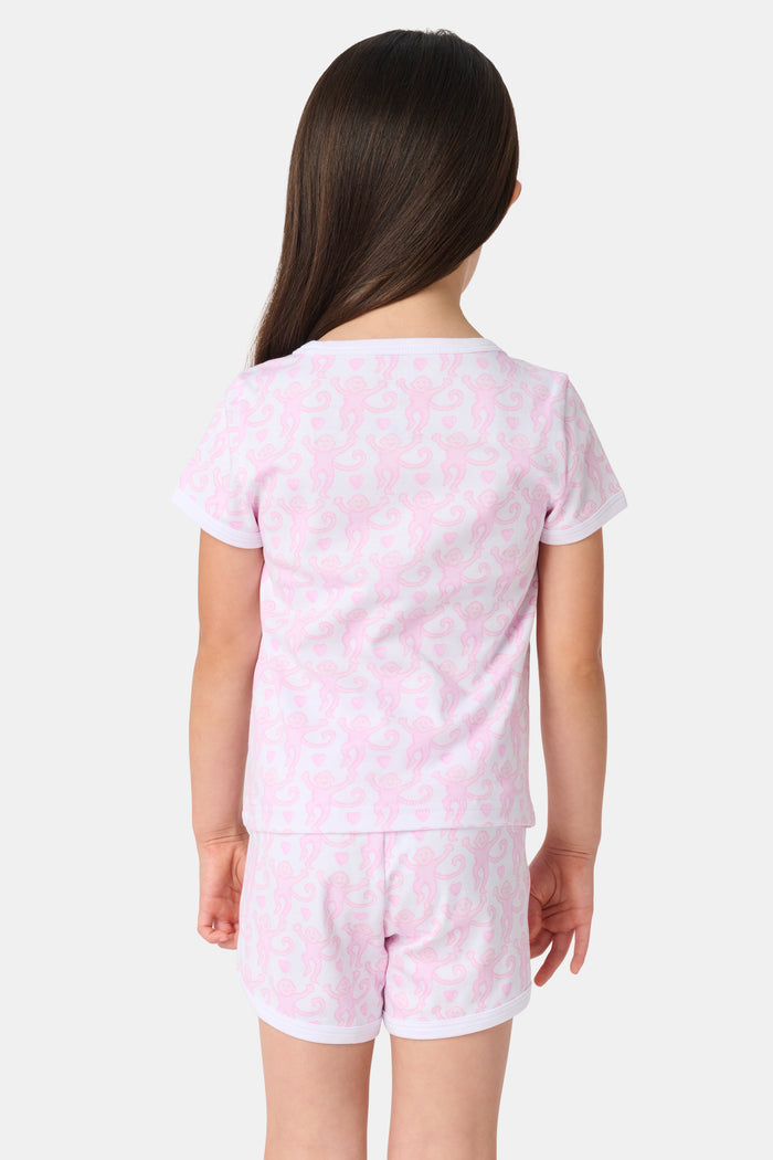 A young girl with straight brown hair wears the Roller Rabbit Kids Lovely Monkey Playa Play Set, a light pink Pima cotton pajama with short sleeves and shorts featuring a subtle monkey pattern as she smiles against a plain white background.