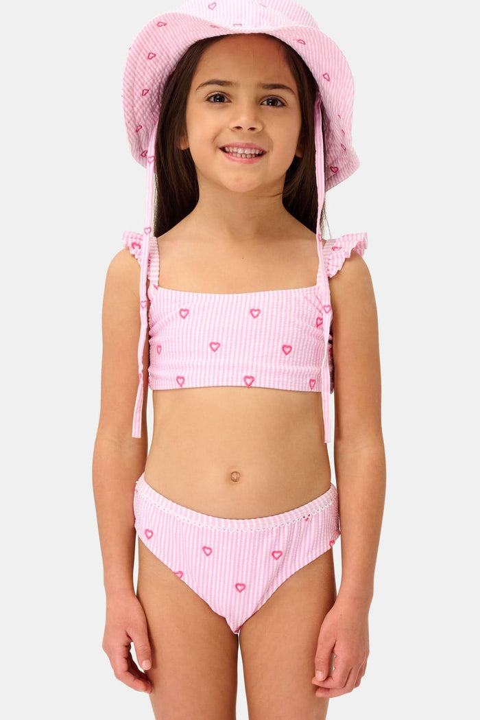 A young girl smiles in the Roller Rabbit Kids Mini Hearts Rory Bikini, featuring pink and white seersucker fabric with heart patterns and ruffle straps, paired with a matching wide-brimmed hat against a plain light background.
