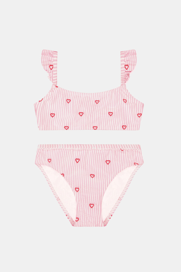 A young girl smiles in the Roller Rabbit Kids Mini Hearts Rory Bikini, featuring pink and white seersucker fabric with heart patterns and ruffle straps, paired with a matching wide-brimmed hat against a plain light background.