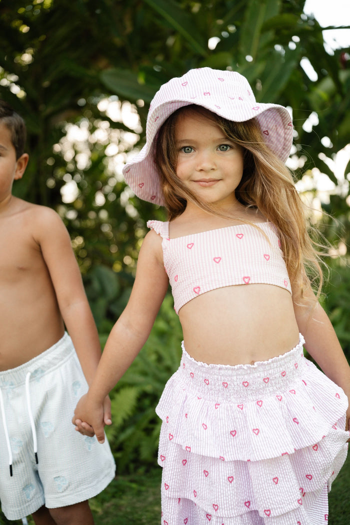 A young girl smiles in the Roller Rabbit Kids Mini Hearts Rory Bikini, featuring pink and white seersucker fabric with heart patterns and ruffle straps, paired with a matching wide-brimmed hat against a plain light background.