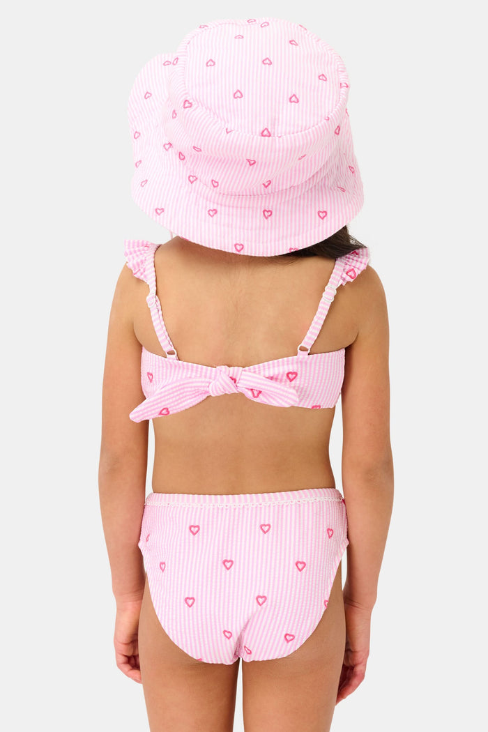 A young girl smiles in the Roller Rabbit Kids Mini Hearts Rory Bikini, featuring pink and white seersucker fabric with heart patterns and ruffle straps, paired with a matching wide-brimmed hat against a plain light background.