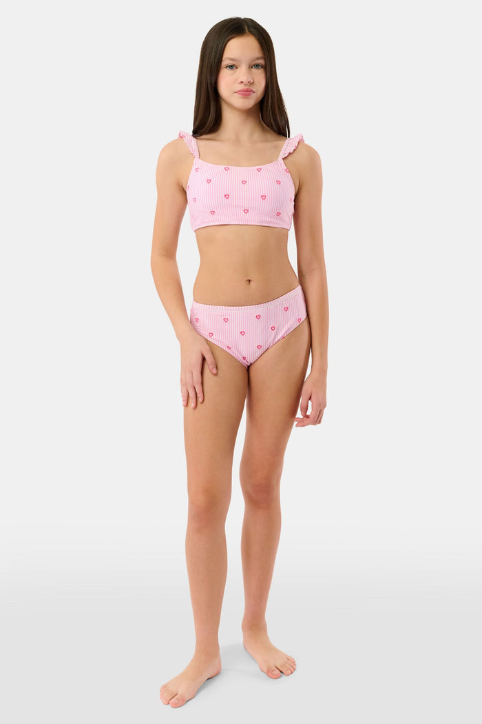 A young girl smiles in the Roller Rabbit Kids Mini Hearts Rory Bikini, featuring pink and white seersucker fabric with heart patterns and ruffle straps, paired with a matching wide-brimmed hat against a plain light background.