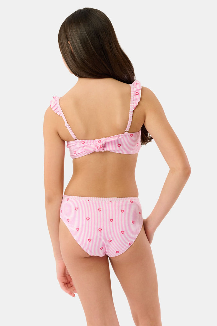 A young girl smiles in the Roller Rabbit Kids Mini Hearts Rory Bikini, featuring pink and white seersucker fabric with heart patterns and ruffle straps, paired with a matching wide-brimmed hat against a plain light background.