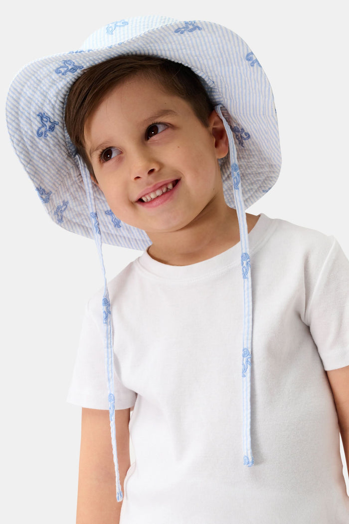 A young child smiles, wearing a cotton white t-shirt and the Roller Rabbit Kids Mini Monkey Bucket Hat—light blue and white stripes with blue floral patterns and chin straps—posing against a light background.