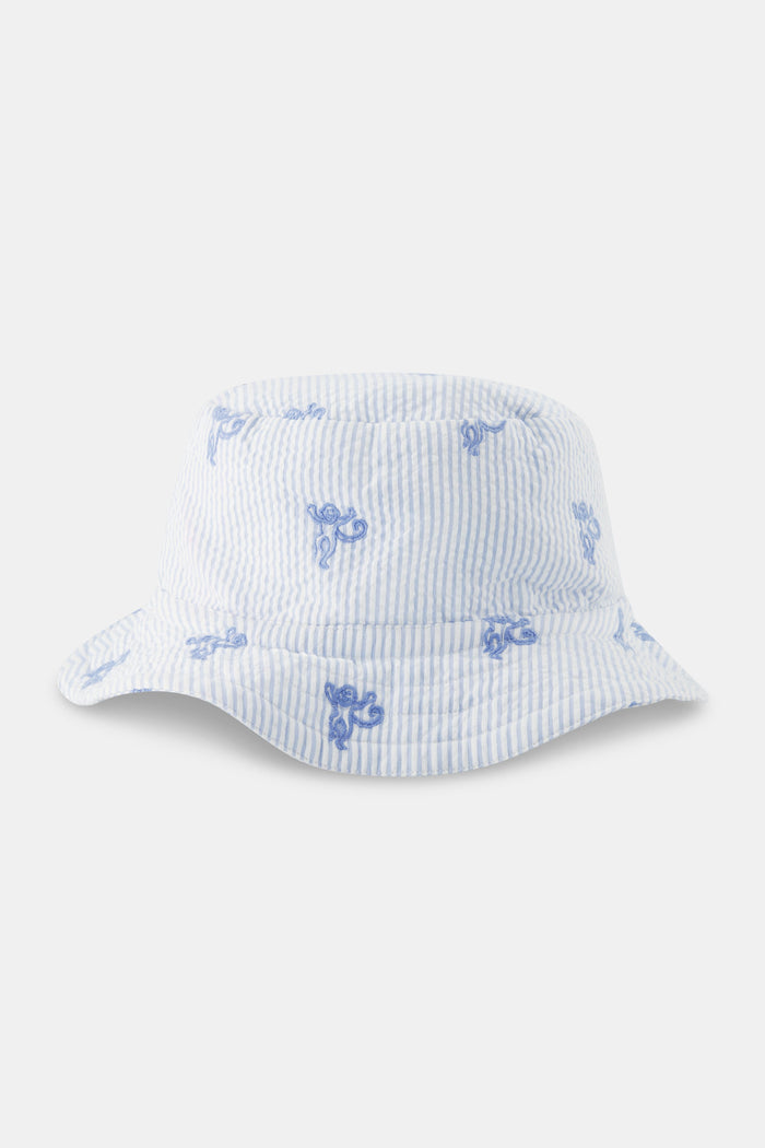 A young child smiles, wearing a cotton white t-shirt and the Roller Rabbit Kids Mini Monkey Bucket Hat—light blue and white stripes with blue floral patterns and chin straps—posing against a light background.