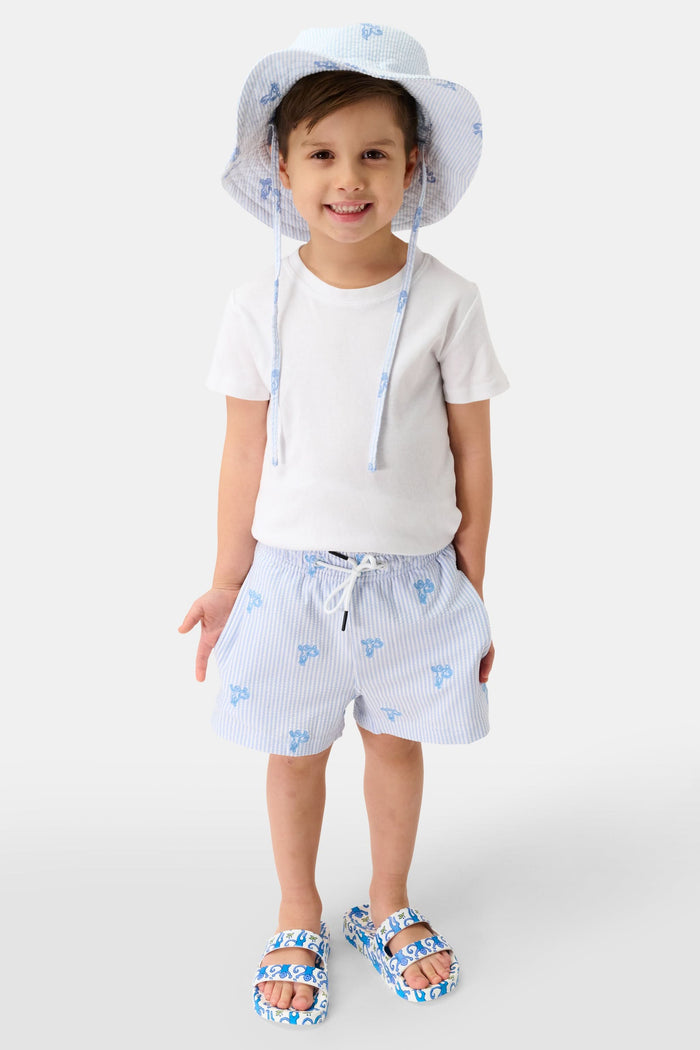 A young child smiles, wearing a cotton white t-shirt and the Roller Rabbit Kids Mini Monkey Bucket Hat—light blue and white stripes with blue floral patterns and chin straps—posing against a light background.