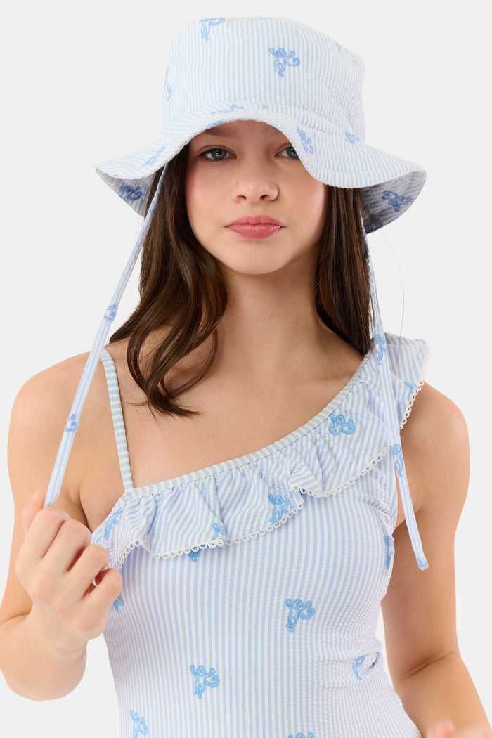 A young child smiles, wearing a cotton white t-shirt and the Roller Rabbit Kids Mini Monkey Bucket Hat—light blue and white stripes with blue floral patterns and chin straps—posing against a light background.
