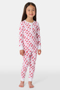 A young child with long brown hair stands barefoot, smiling and wearing the Roller Rabbit Kids Moby Pajamas made from soft Pima cotton, featuring a pink and red mitten pattern, against a plain light background.