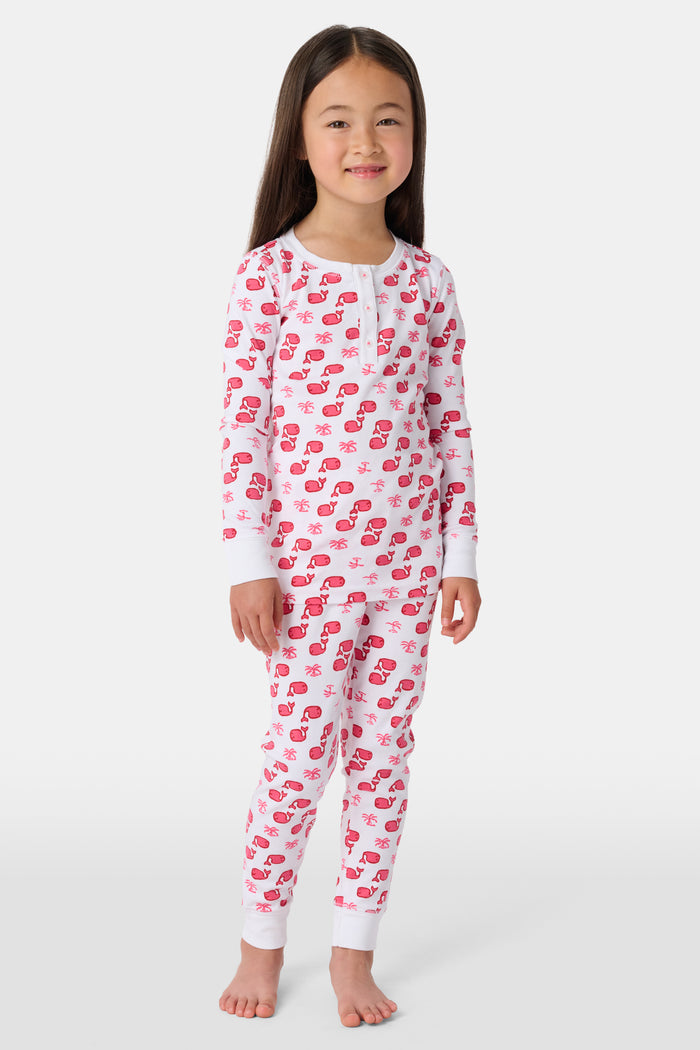 A young child with long brown hair stands barefoot, smiling and wearing the Roller Rabbit Kids Moby Pajamas made from soft Pima cotton, featuring a pink and red mitten pattern, against a plain light background.