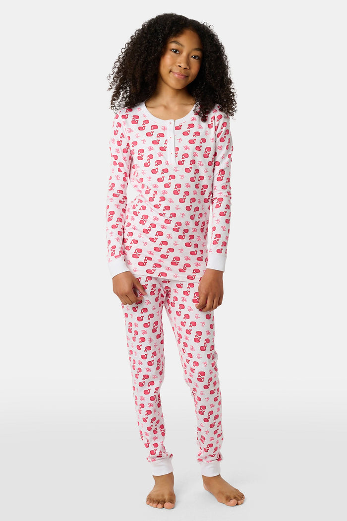 A young child with long brown hair stands barefoot, smiling and wearing the Roller Rabbit Kids Moby Pajamas made from soft Pima cotton, featuring a pink and red mitten pattern, against a plain light background.