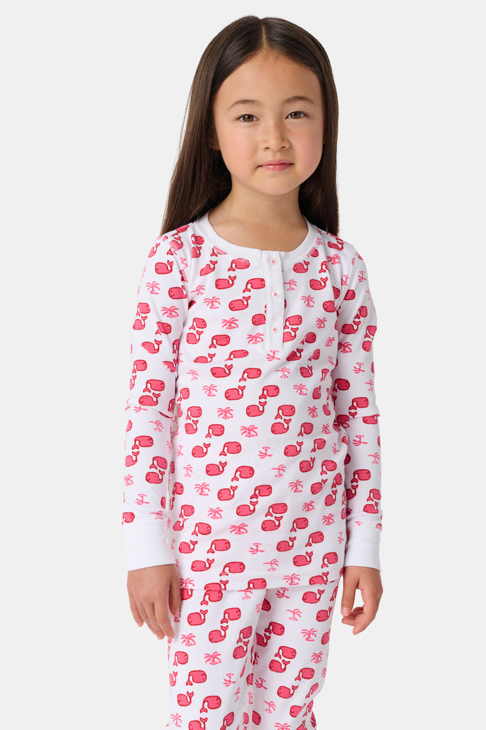 A young child with long brown hair stands barefoot, smiling and wearing the Roller Rabbit Kids Moby Pajamas made from soft Pima cotton, featuring a pink and red mitten pattern, against a plain light background.
