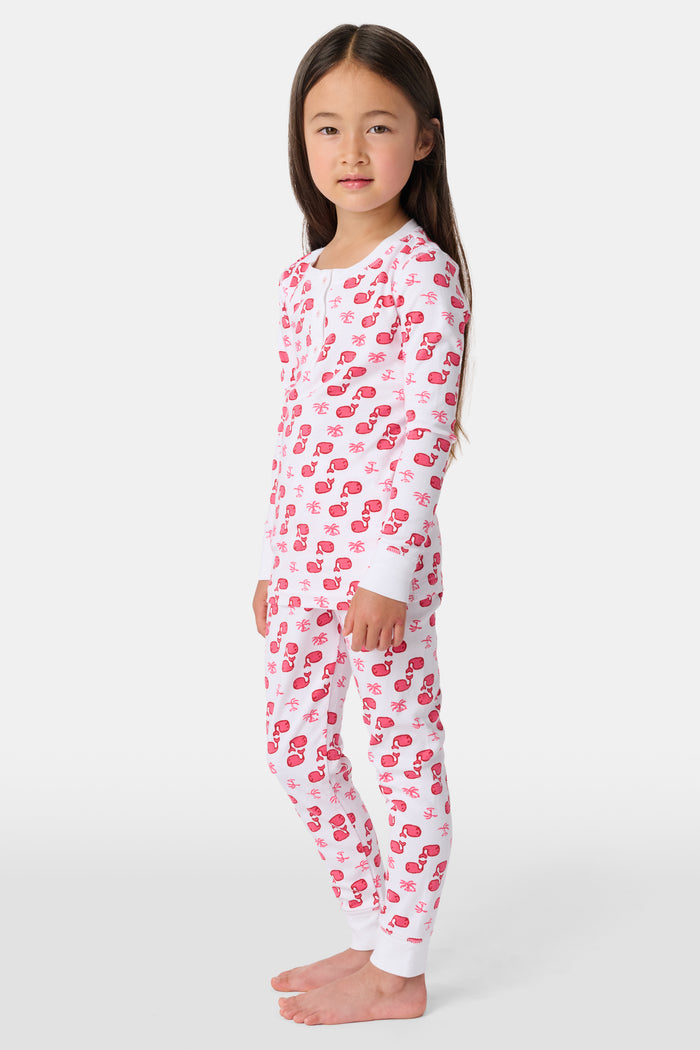 A young child with long brown hair stands barefoot, smiling and wearing the Roller Rabbit Kids Moby Pajamas made from soft Pima cotton, featuring a pink and red mitten pattern, against a plain light background.