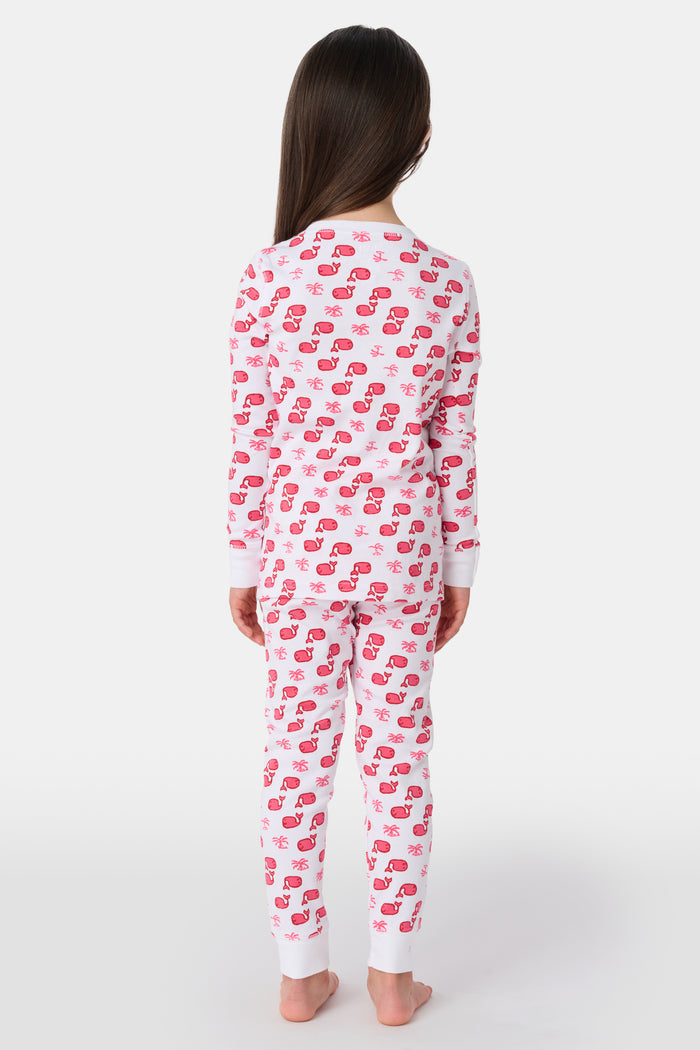 A young child with long brown hair stands barefoot, smiling and wearing the Roller Rabbit Kids Moby Pajamas made from soft Pima cotton, featuring a pink and red mitten pattern, against a plain light background.