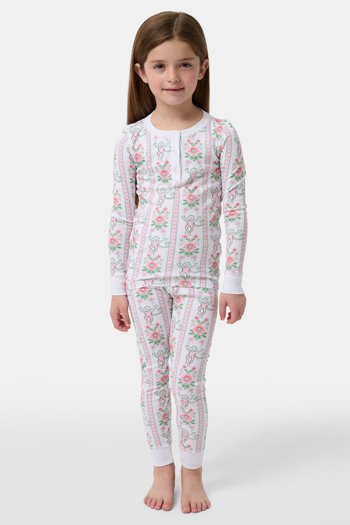 A young girl stands barefoot in Roller Rabbit Kids Monkey Block Stripe Pajamas—matching long-sleeve top and pants with playful monkey block stripes—smiling against a plain light gray background.