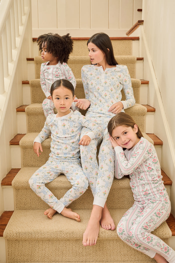 A young girl stands barefoot in Roller Rabbit Kids Monkey Block Stripe Pajamas—matching long-sleeve top and pants with playful monkey block stripes—smiling against a plain light gray background.