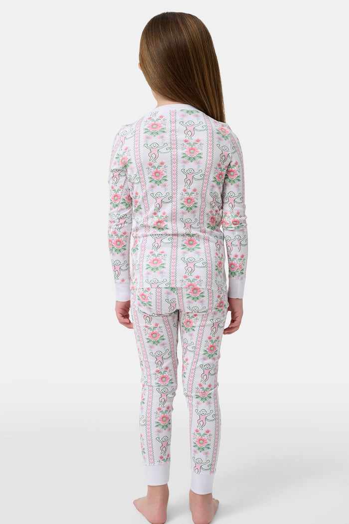 A young girl stands barefoot in Roller Rabbit Kids Monkey Block Stripe Pajamas—matching long-sleeve top and pants with playful monkey block stripes—smiling against a plain light gray background.