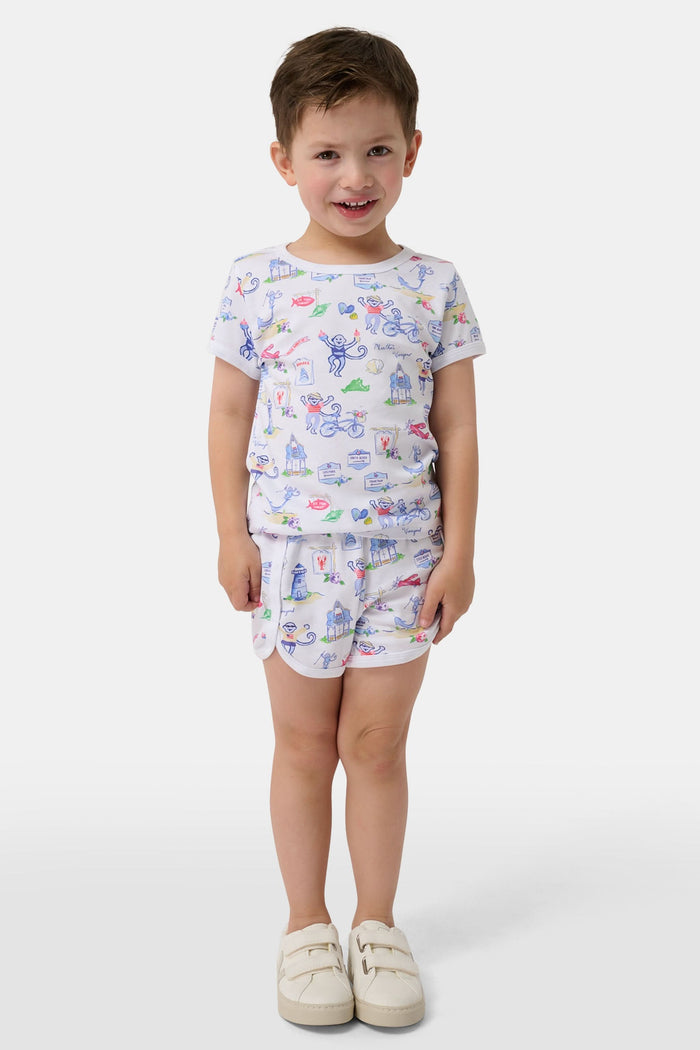A young child stands barefoot, smiling and facing forward, wearing Roller Rabbit Kids Monkey On The Vineyard Pajamas made of soft Pima cotton with colorful cartoon patterns of animals, vehicles, and buildings. The pajamas feature buttons at the neckline.