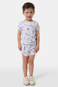 A young child stands barefoot, smiling and facing forward, wearing Roller Rabbit Kids Monkey On The Vineyard Pajamas made of soft Pima cotton with colorful cartoon patterns of animals, vehicles, and buildings. The pajamas feature buttons at the neckline.